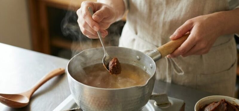味噌汁の調理シーン。コンロの火を止めた後、湯気が立つ鍋にスプーンで味噌を溶き入れている様子。