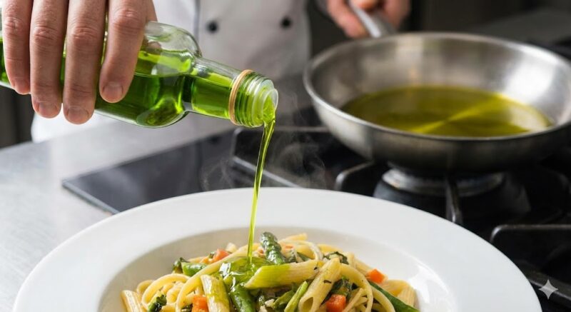 出来上がった料理に、鮮やかな緑色のエキストラバージンオリーブオイルを仕上げに回しかけている様子。背景のフライパンからは煙が出ておらず、適切な中温調理であったことがわかる